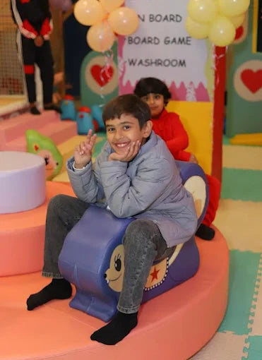 A young boy smiles while sitting on a purple toy at a play area, with another child in the background.