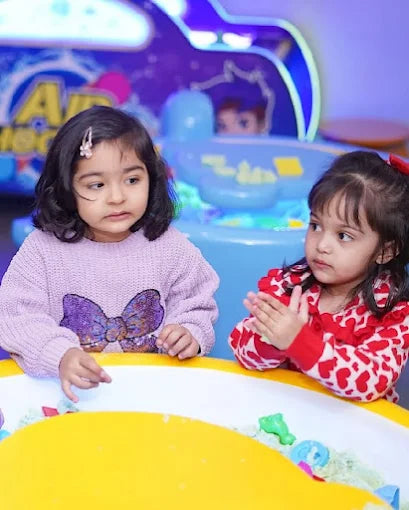 Two young girls play with kinetic sand at a yellow table in front of a colorful arcade game.