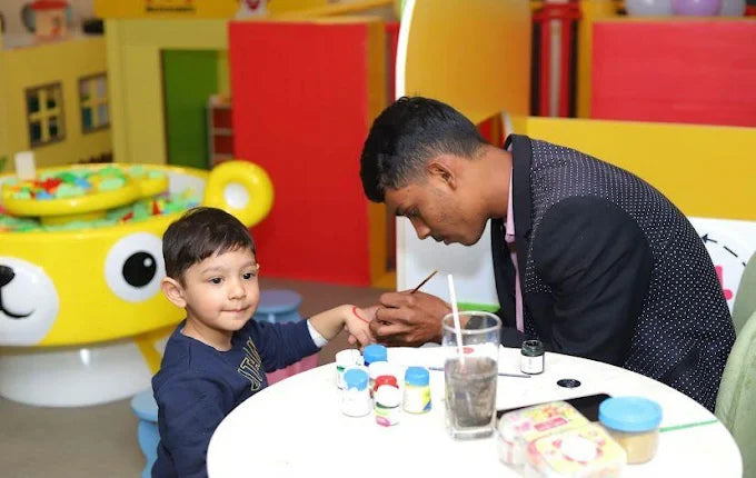 A man paints a child's hand at a table with art supplies.