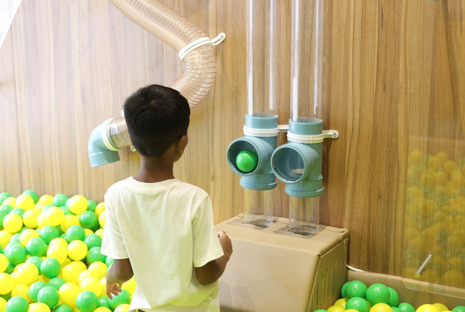 A young boy plays with balls in a ball pit, interacting with a tube system on a wooden wall.