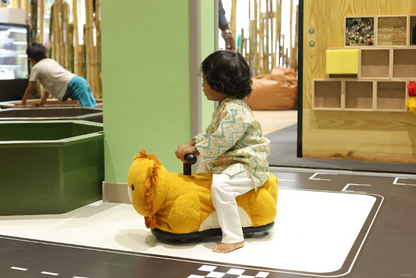 A young child rides a stuffed lion toy in a play area with a green wall and wooden shelves.