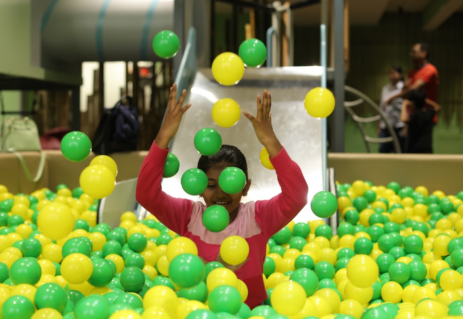 A young girl joyfully plays in a ball pit, tossing green and yellow balls in the air.