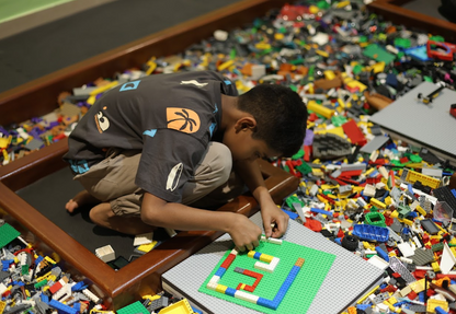 A young boy kneels and plays with LEGOs in a large bin filled with colorful bricks. He is building on a green baseplate.