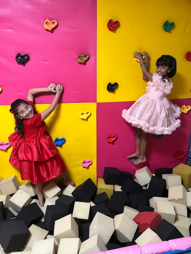 Two children in colorful dresses playing in a foam pit with a multicolored wall.