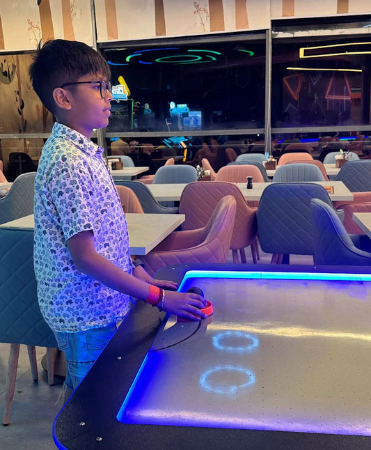 A young boy plays air hockey in a brightly lit arcade with colorful chairs in the background.