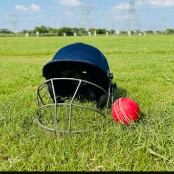 Cricketer's helmet and red ball on a grassy field with power lines in the background