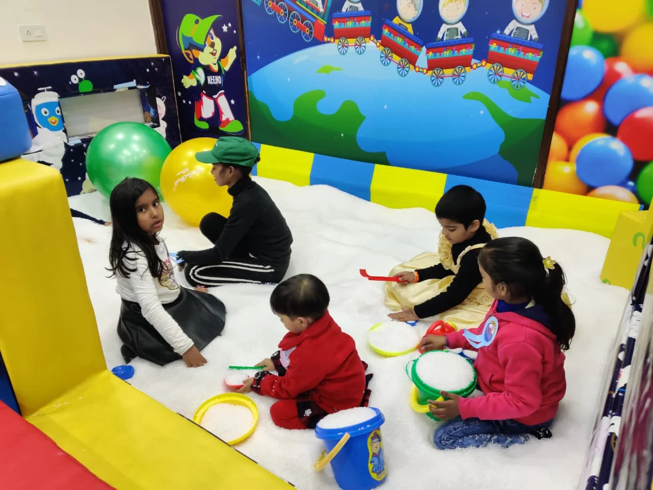 Children playing with toys in a colorful playroom with a large screen displaying cartoon characters.