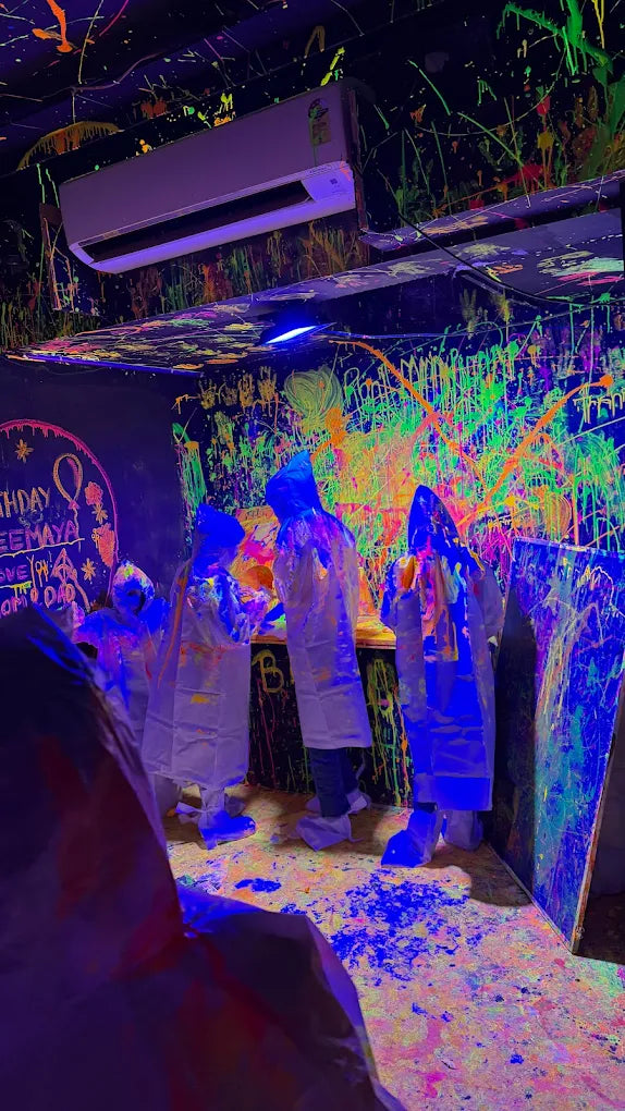 People in protective gear engage in a paint-splattering activity in a neon-lit room. The walls and floor are covered in vibrant, colorful paint splatters.