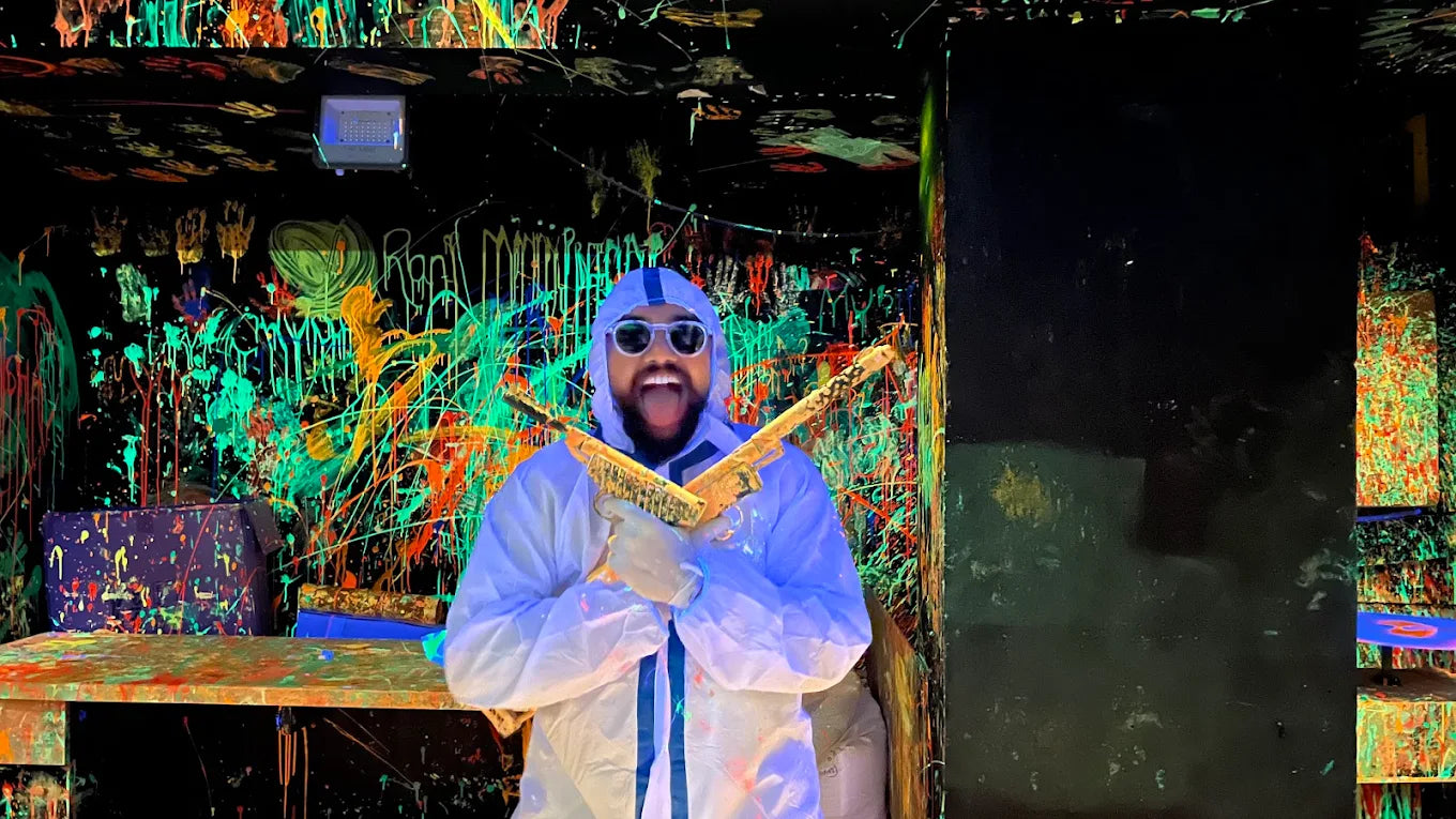 A person in a white suit holds two golden guns in a room splattered with neon paint.