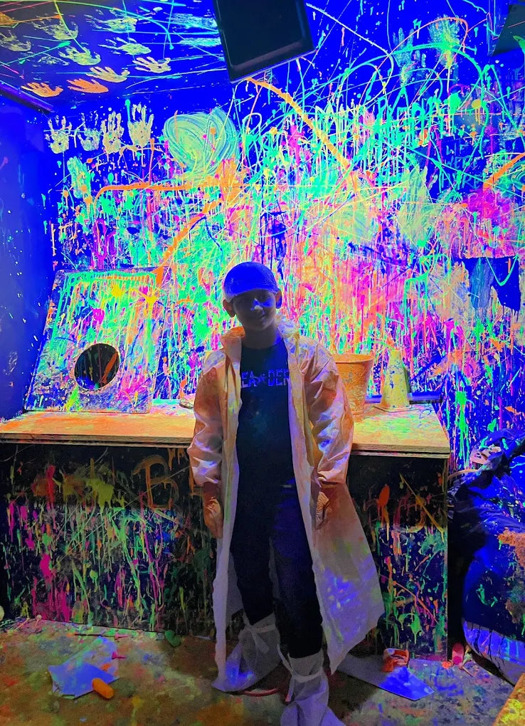A child in a lab coat stands in a room covered in neon paint splatters. The walls and ceiling are a vibrant canvas of neon colors.