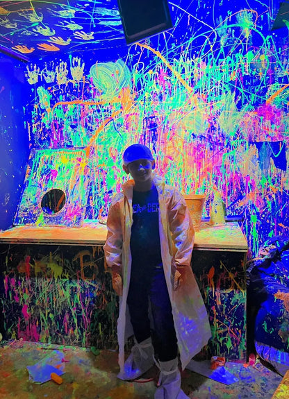 A child in a lab coat stands in a room covered in neon paint splatters. The walls and ceiling are a vibrant canvas of neon colors.
