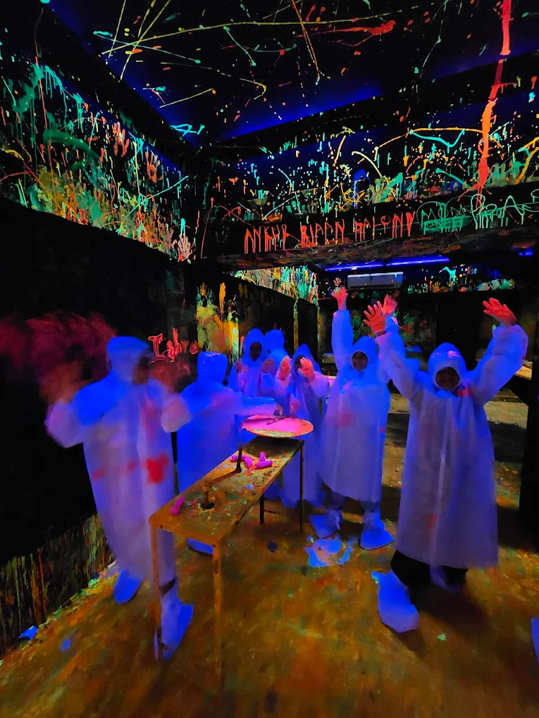 People in protective suits in a dark room splattered with neon paint, with some raising their hands in excitement.