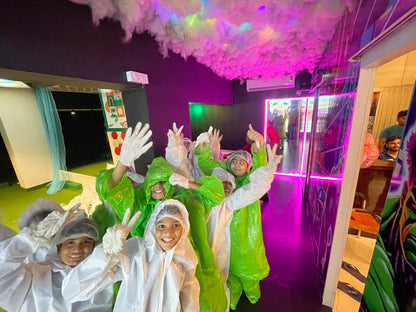 Children in protective suits and gloves pose for a photo in a colorful, neon-lit room with a cloud-like ceiling.