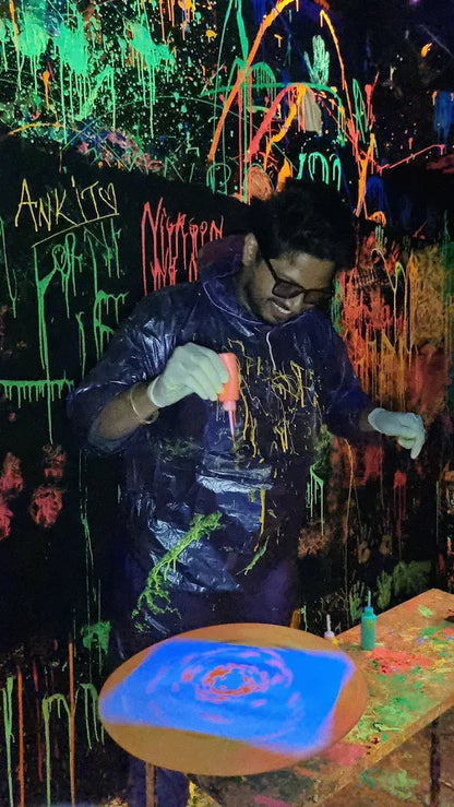 A young person in a paint-splattered poncho creates art with neon paint in a blacklight room.