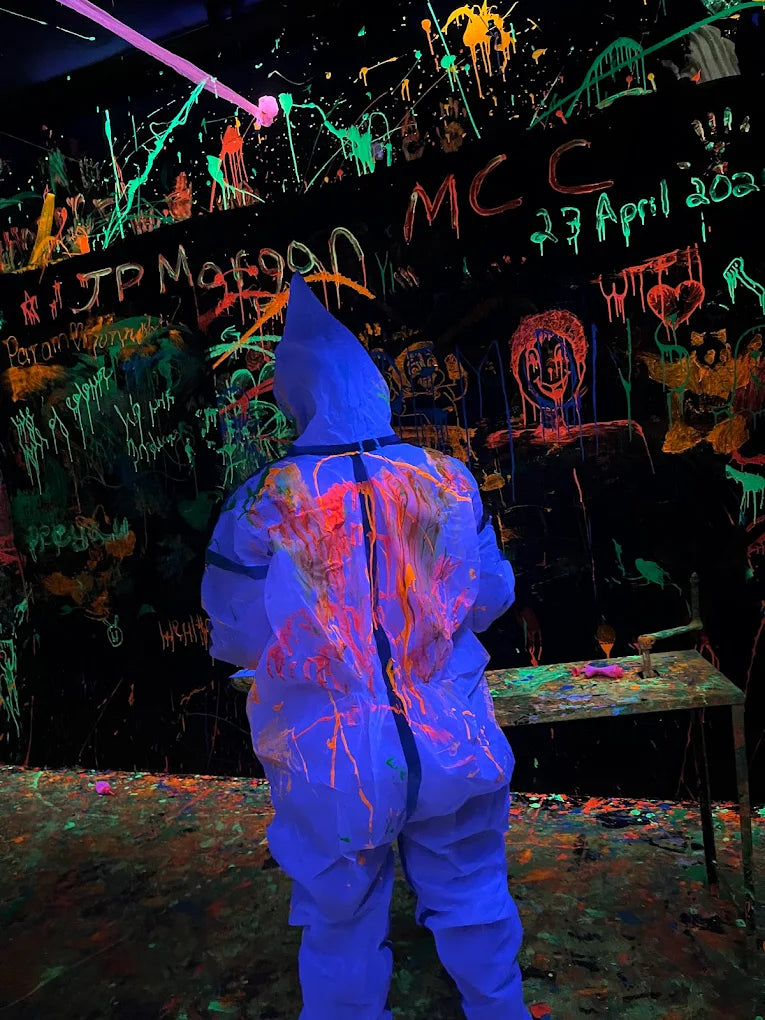 A person in a blue protective suit covered in neon paint splatters stands in a dark room with graffiti.