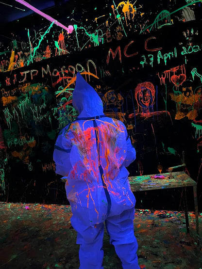 A person in a blue protective suit covered in neon paint splatters stands in a dark room with graffiti.