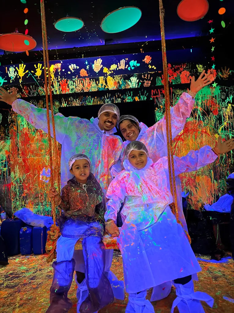 A family in protective suits poses in a neon-lit room covered in colorful paint splatters.