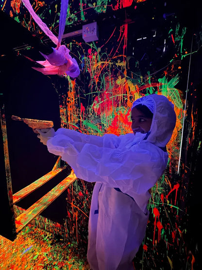 A child in a white suit holds a paint gun in a neon-splattered room.