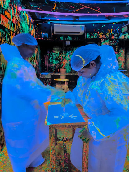 Two people in protective suits paint a stick figure on a blue surface in a neon-splattered room.