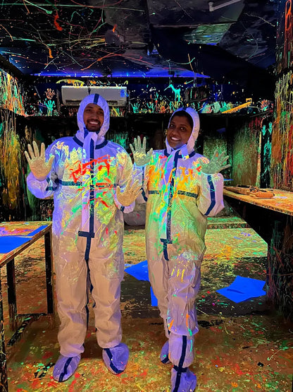 Two adults in protective suits covered in neon paint stand in a dark room splattered with colorful paint.