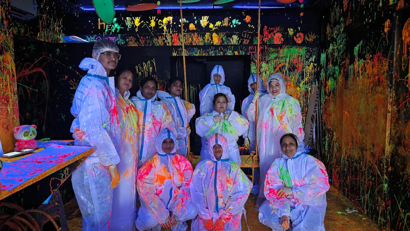 A group of adults in protective suits are covered in neon paint in a dark, splatter-painted room.
