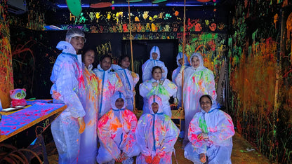 A group of adults in protective suits are covered in neon paint in a dark, splatter-painted room.