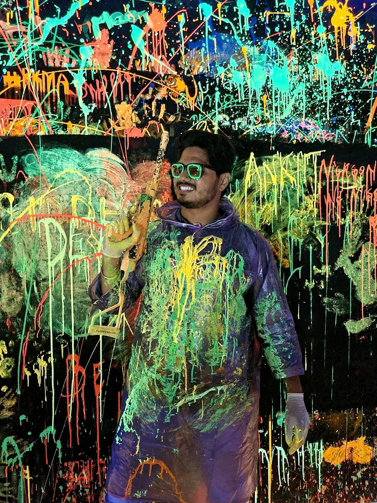 A young man in neon glasses and a paint-splattered poncho stands in front of a black wall covered in colorful paint splatters.