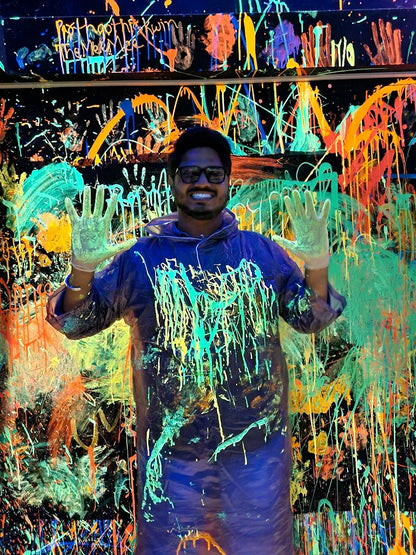 A young man in a paint-splattered smock and gloves smiles, holding up his hands in front of a black wall covered in neon paint splatters.