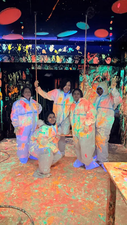 Adults in paint-splattered suits pose in a neon-lit room covered in handprints and paint splatters.