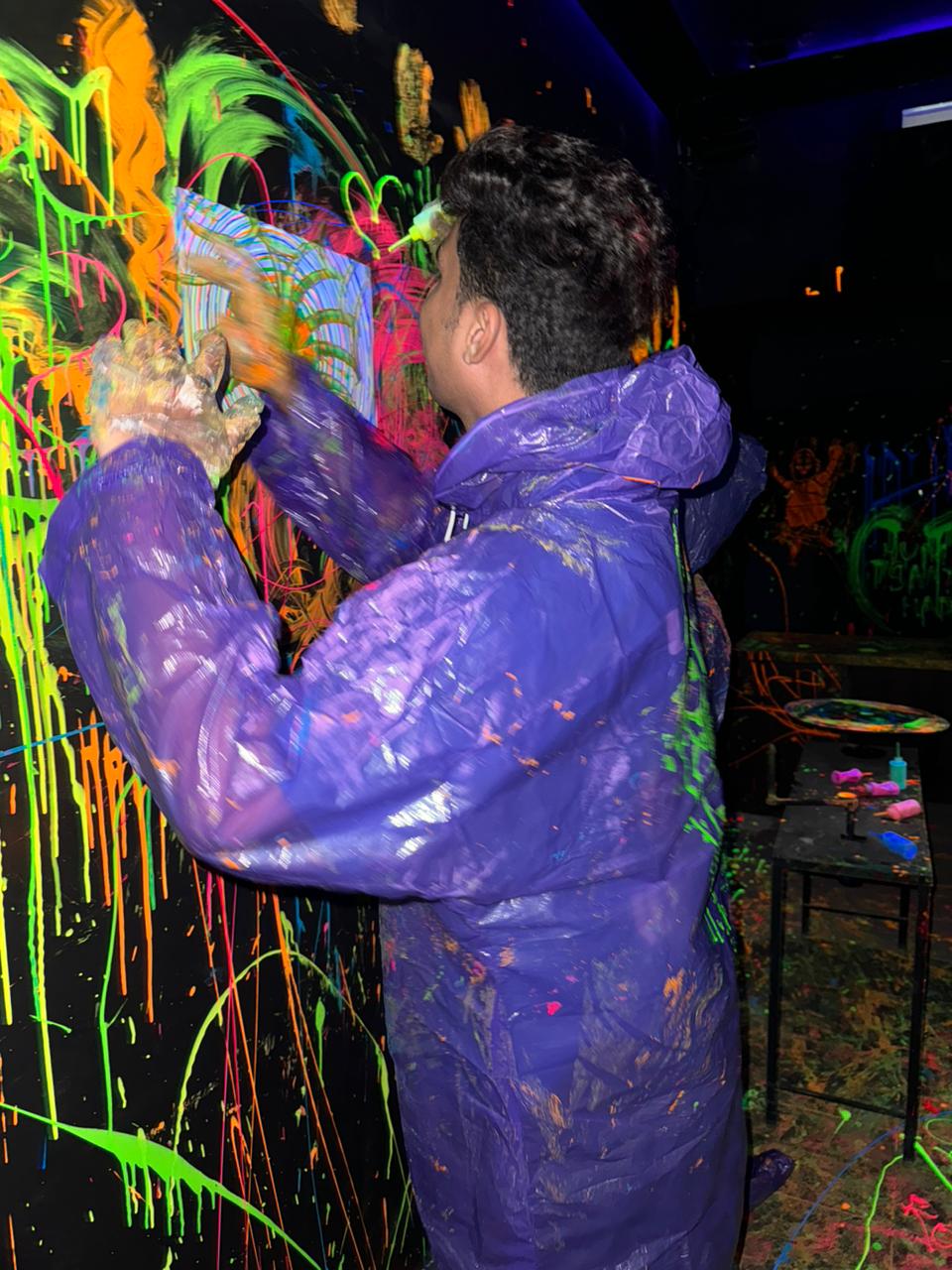 A young person in a purple poncho splatters neon paint on a black wall in a dark room.
