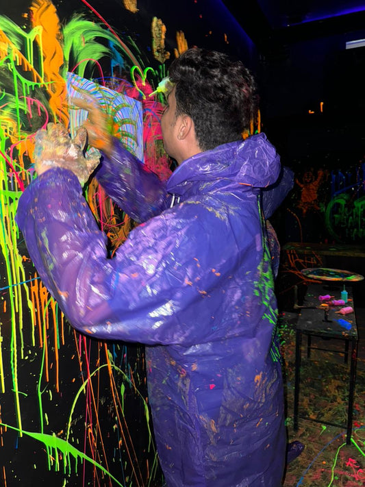 A young person in a purple poncho splatters neon paint on a black wall in a dark room.