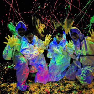 Four young people in protective suits covered in neon paint, posing in a dark, paint-splattered room.