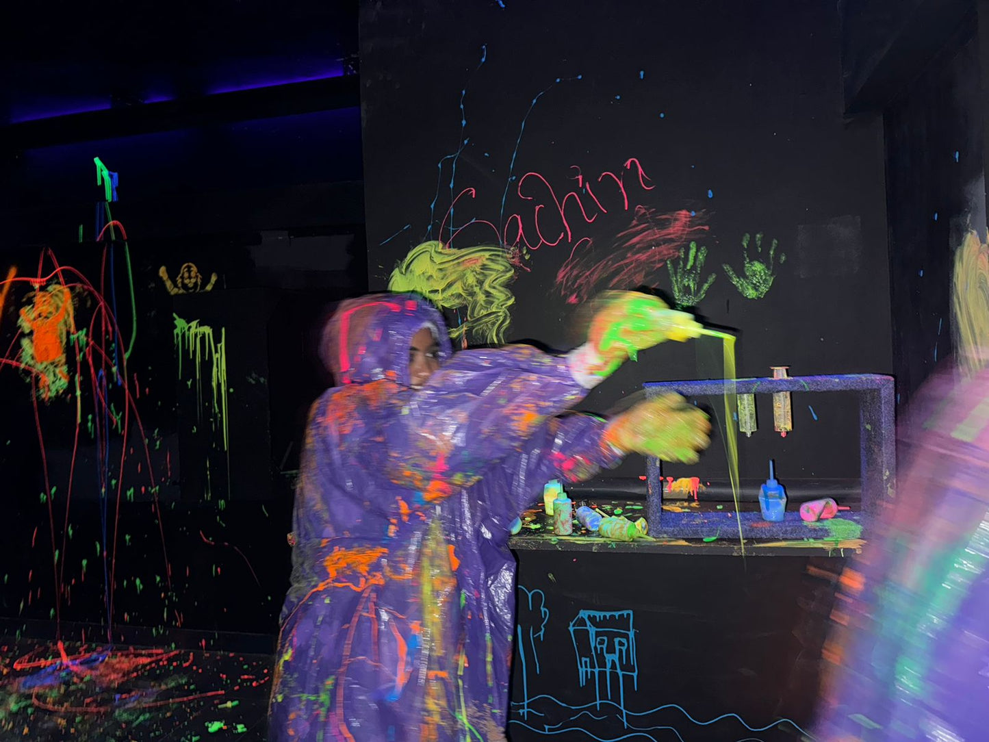A person in a paint-splattered poncho throws neon paint in a dark room with glowing art.