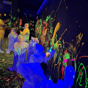 People in protective suits splatter neon paint on a black wall in a dark, glow-in-the-dark room.