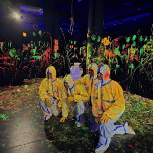 People in protective suits in a dark room splattered with neon paint.