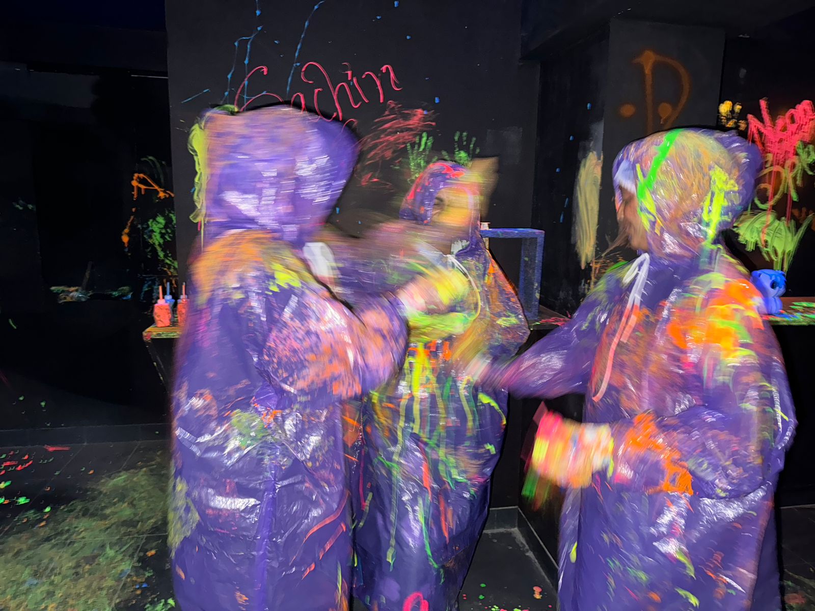 People in purple ponchos covered in neon paint splatters in a dark, graffiti-covered room.
