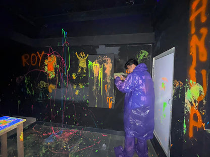 A young person in a purple poncho paints on a black wall covered in neon splatters and drawings.