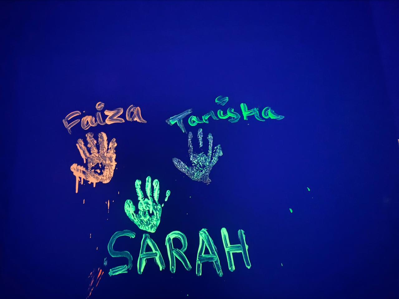 Children's names and handprints glow under blacklight on a dark blue wall.