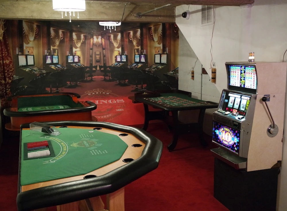 A casino-themed room with slot machines, a roulette table, and a blackjack table.