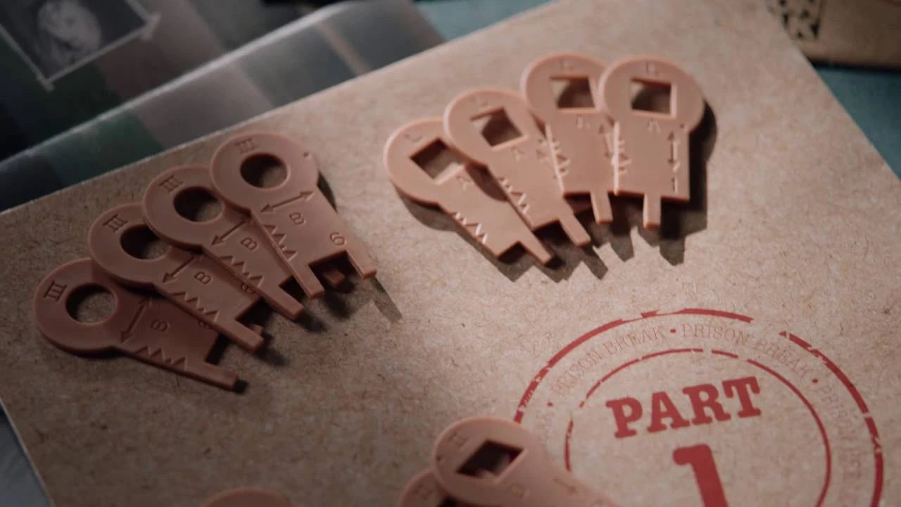 Plastic game pieces resembling keys are scattered on a cardboard surface with a "Prison Break Part 1" stamp.