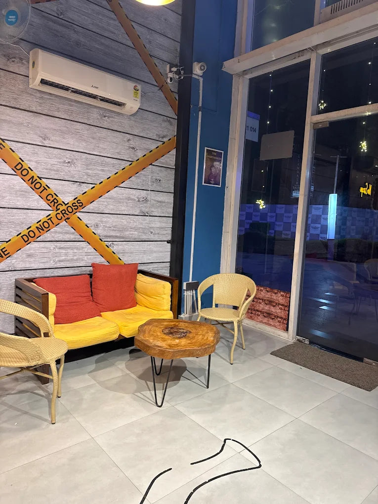 A modern seating area with a yellow couch, chairs, and a wooden table. The wall has "DO NOT CROSS" tape.