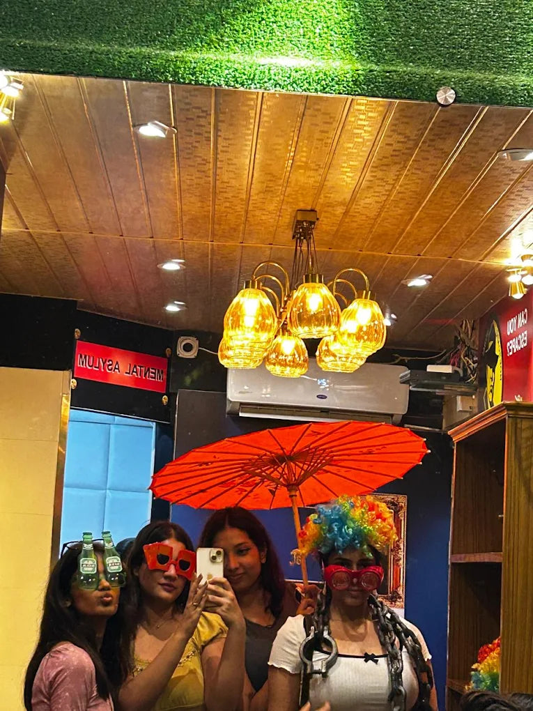 Four young women in a photo booth with props, including a colorful wig and novelty glasses.