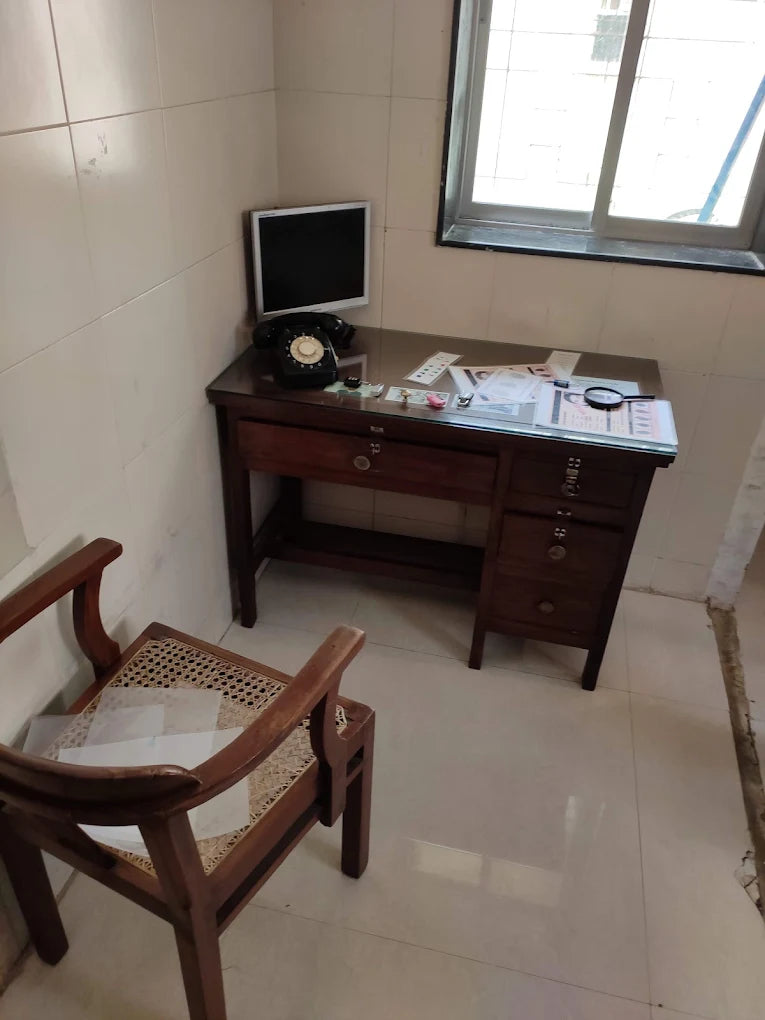 A vintage desk with a rotary phone and a magnifying glass sits in a room with tiled walls and floor. A wooden chair with a woven seat is in the foreground.