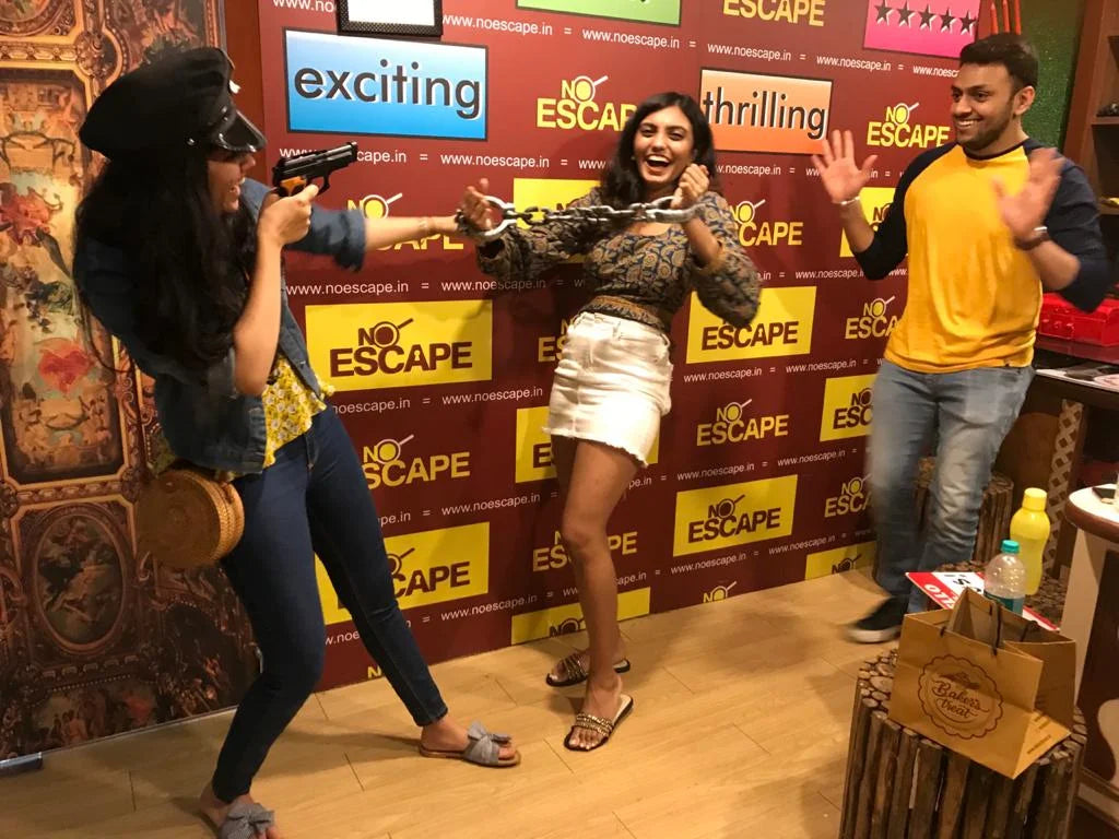 Young adults play an escape room game, one holding a toy gun and chains, the other waving.