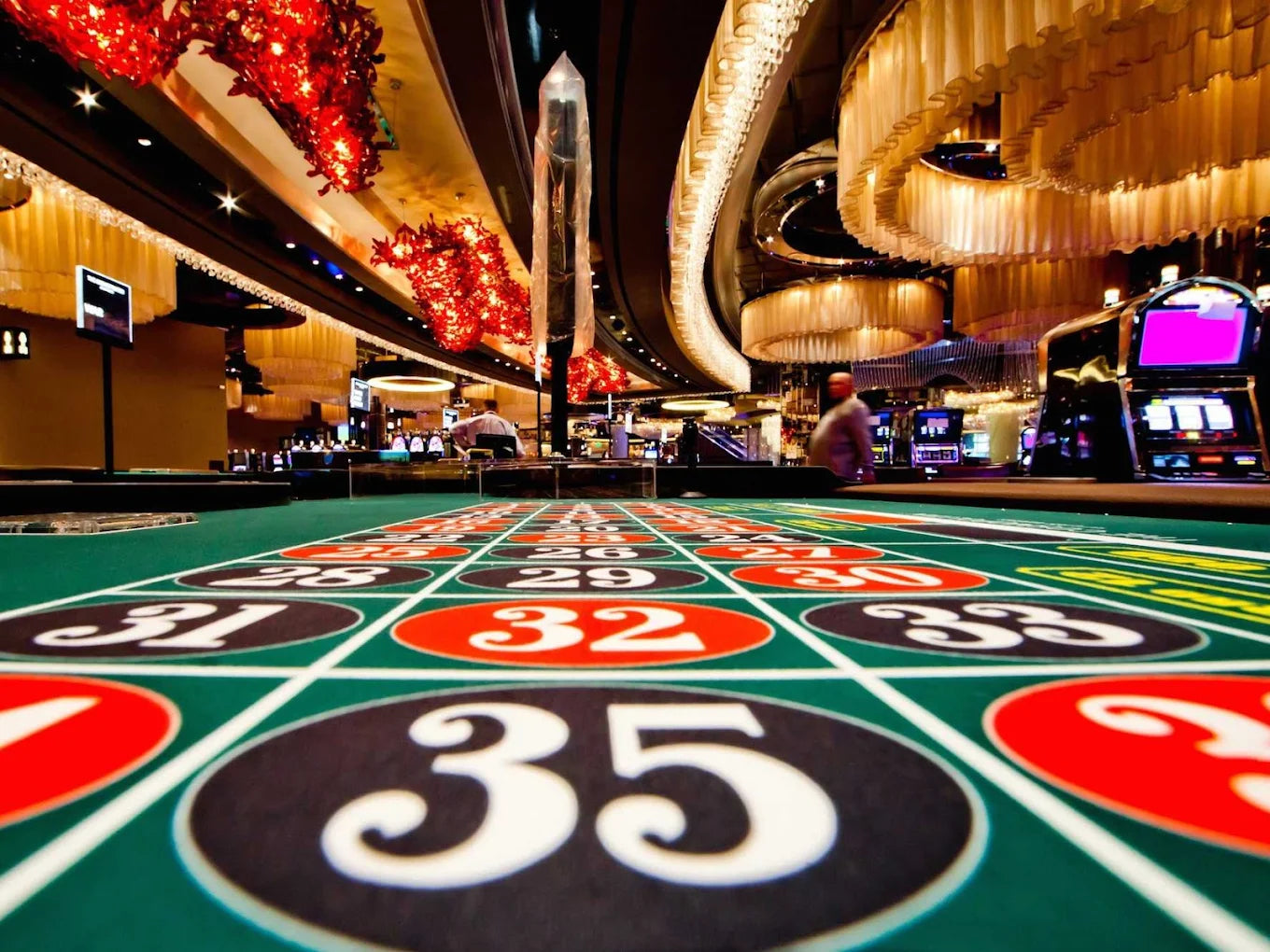 A roulette table in a casino with slot machines and chandeliers in the background.