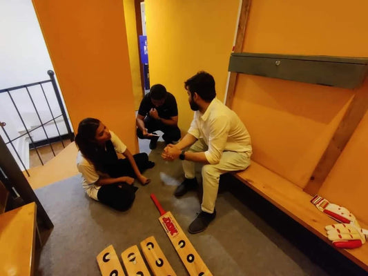 Young adults sit on the floor and a bench in a room with orange walls, with cricket equipment nearby.