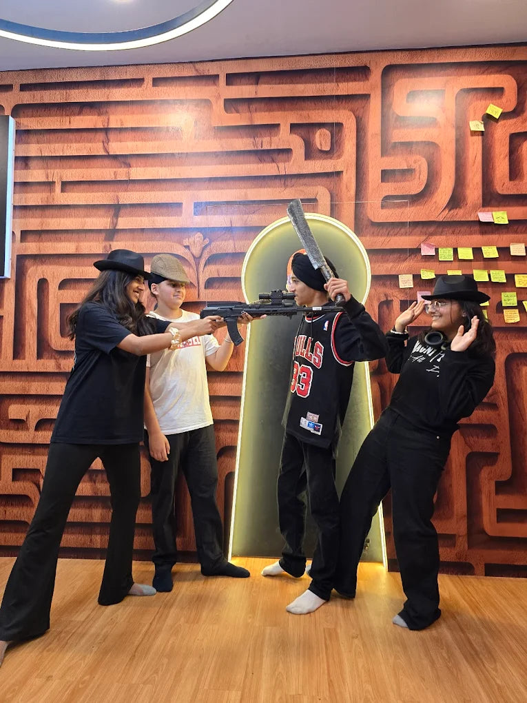 Four young people in hats pose with toy weapons in front of a maze-like wall.