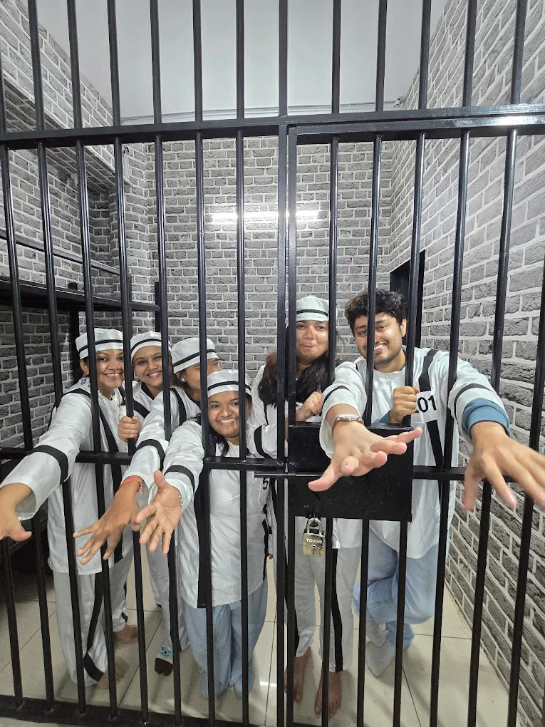 A group of young adults in striped prison outfits pose behind bars, smiling and reaching out.