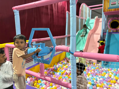 A young boy plays with a toy gun in a colorful ball pit at an indoor playground. Other children are visible on slides in the background.