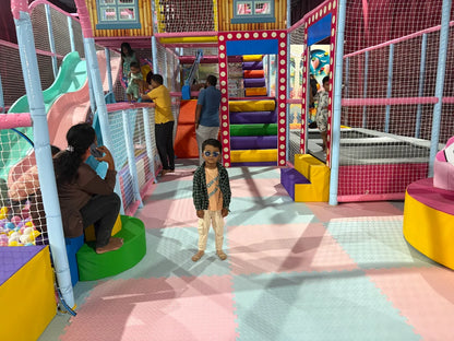 A young boy stands in a colorful indoor playground while a woman talks on her phone in the background.